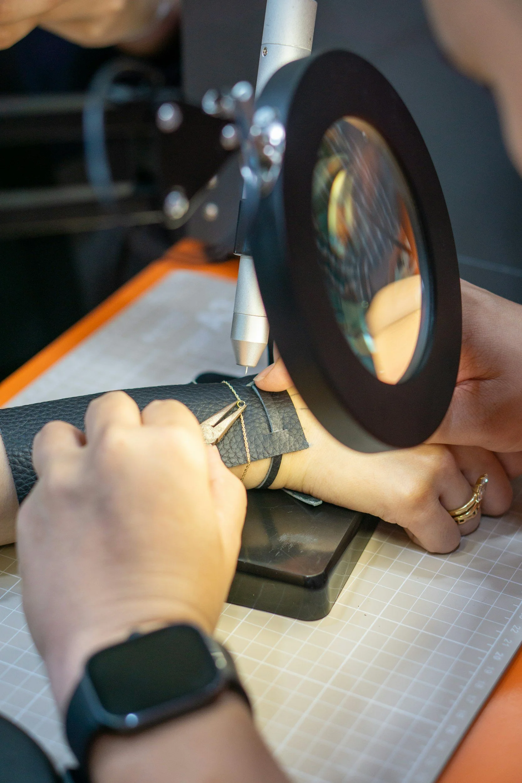Close-up of hands working on jewelry with a loupe, using precision tools on a black textured material, on a grid-lined workspace.