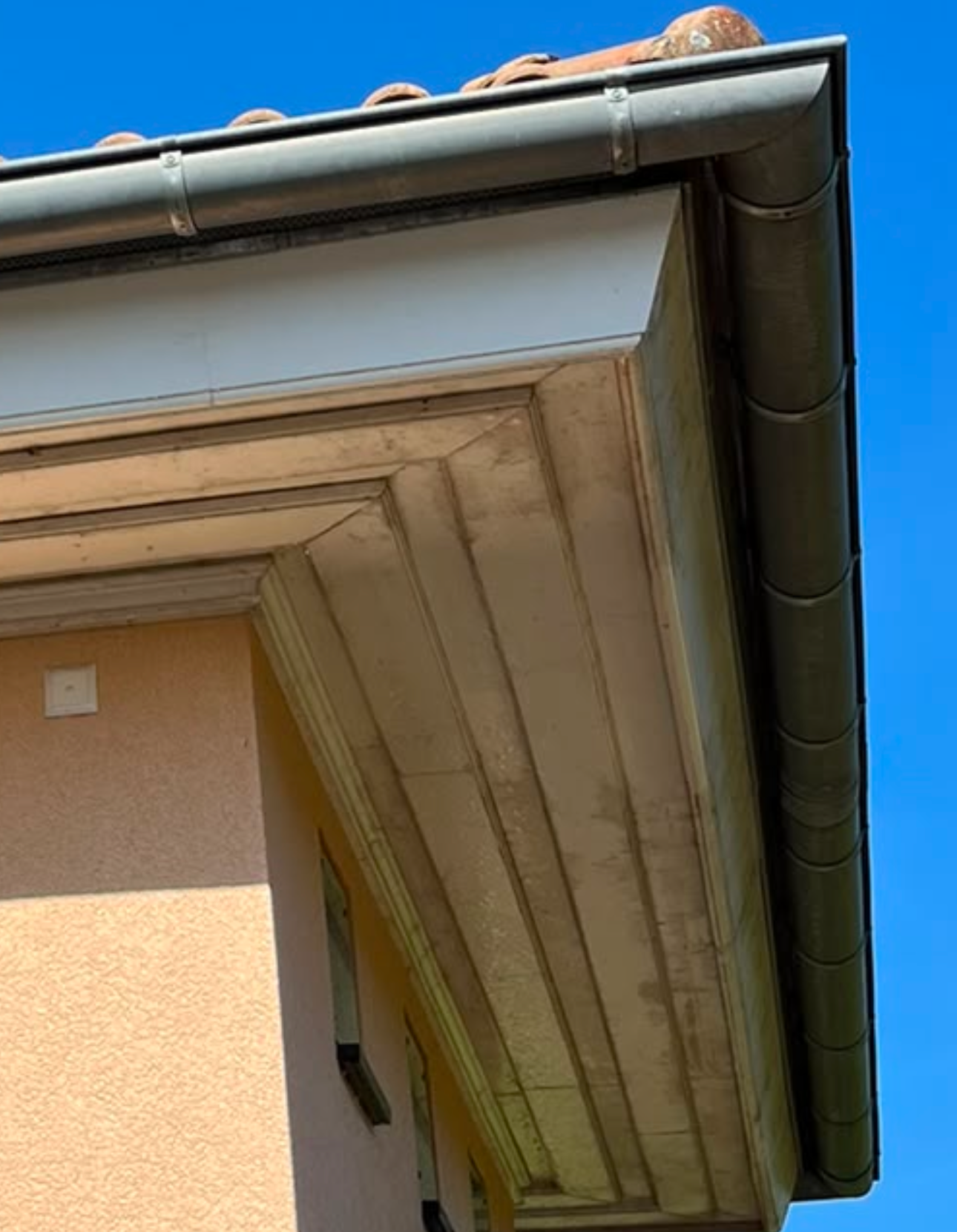Der Blick auf die Ecke eines Gebäudes mit Holzbalken, einer weißen Außenwand, einem orangefarbenen Bereich und einem Dach mit Regenrinnen gegen einen blauen Himmel.