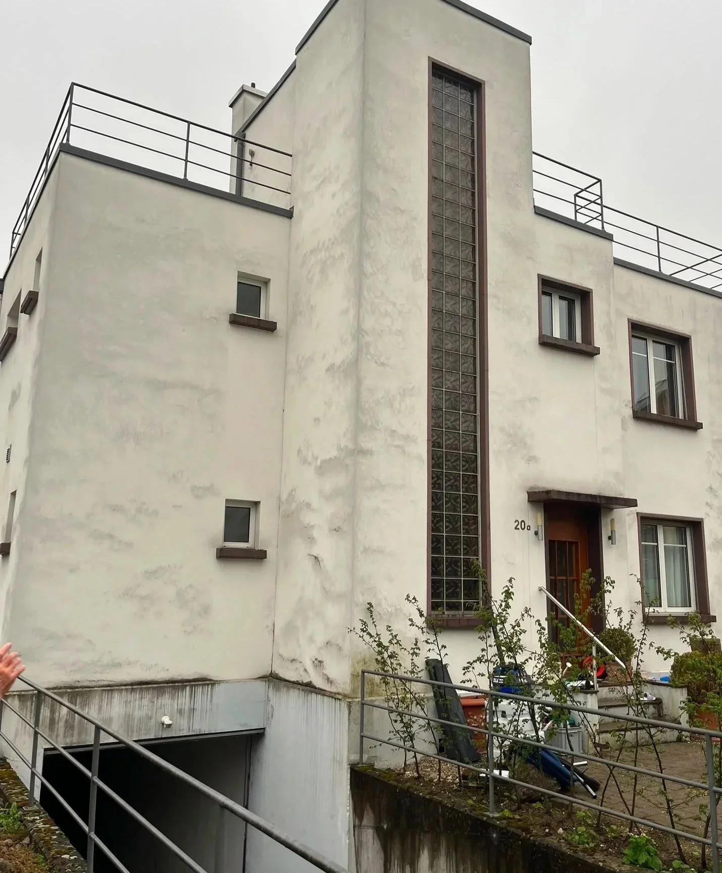 Mehrstöckiges modernes Haus mit weißen Wänden, mehreren Fenstern und einem vertikalen Glasblockfenster. Die Terrasse auf dem Dach hat eine Metallbrüstung. Der Eingang befindet sich im unteren rechten Bereich, daneben eine kleine Treppe und ein Garten mit Pflanzen.