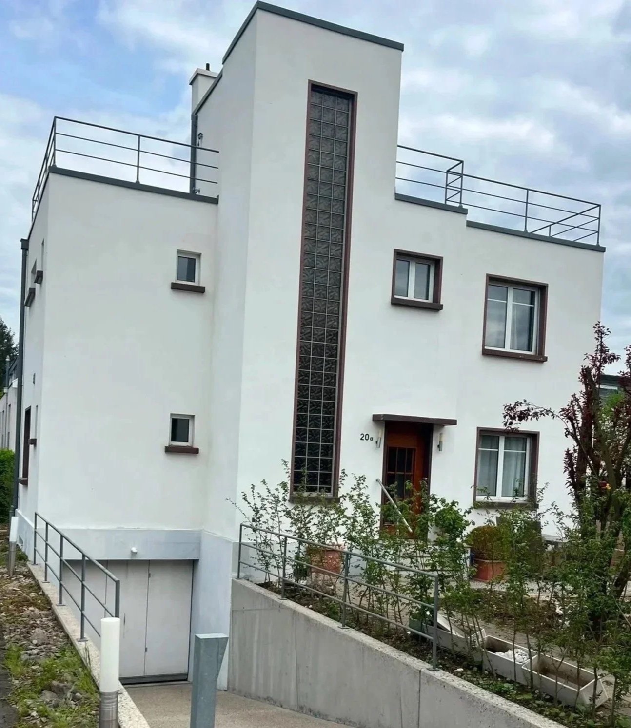 Neues modernes weißes Mehrfamilienhaus mit mehreren Fenstern, Dachterrassen und vertikalem Glasblockfenster an der Fassade, umgeben von kleineren Büschen und Pflanzen, mit einem gepflasterten Weg und einem Rolltor im Vordergrund.
