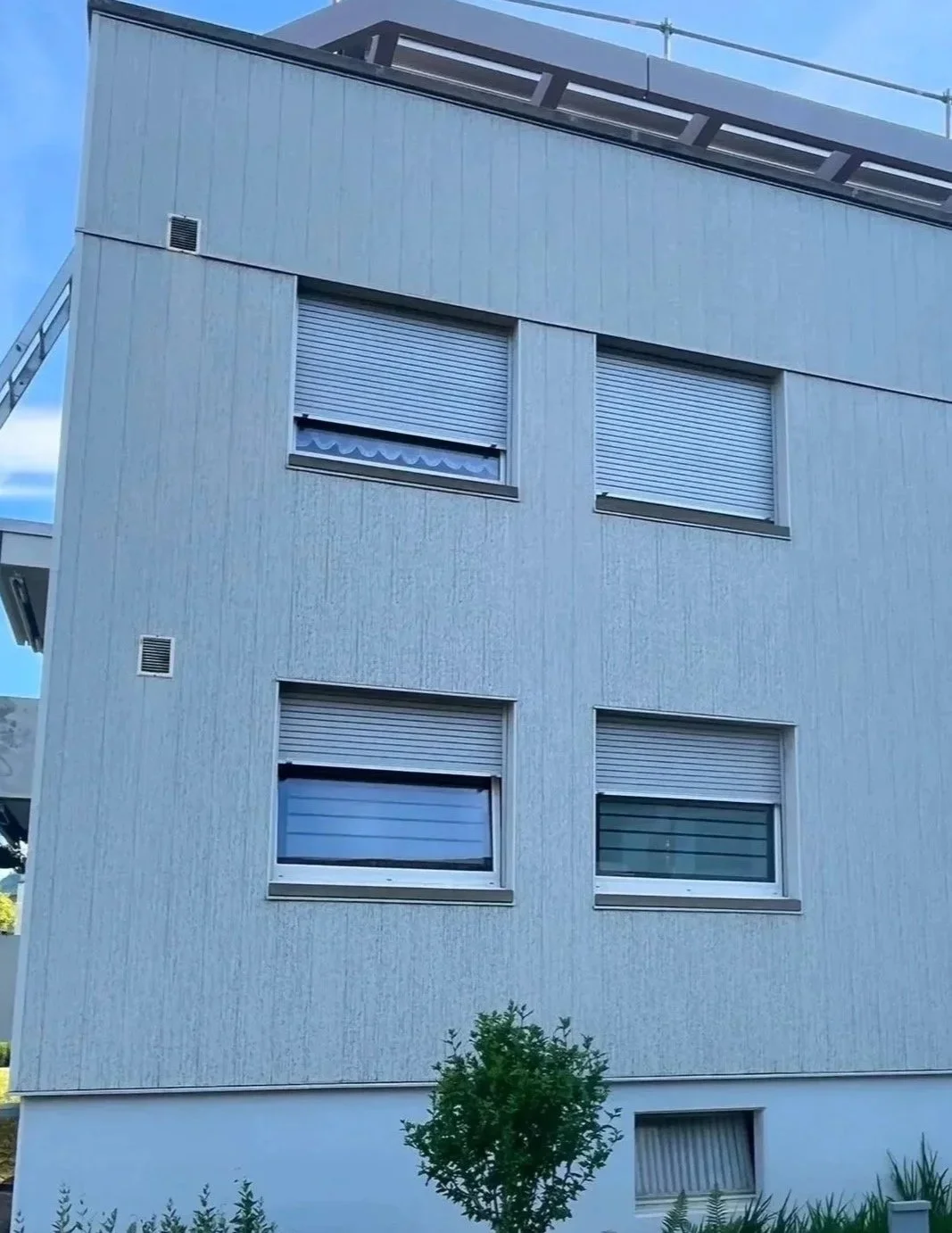 Mehrstöckiges modernes Gebäude mit weißen Wandflächen, mehreren Fenstern mit Rollläden, kleine Bäume und Pflanzen im Vordergrund.