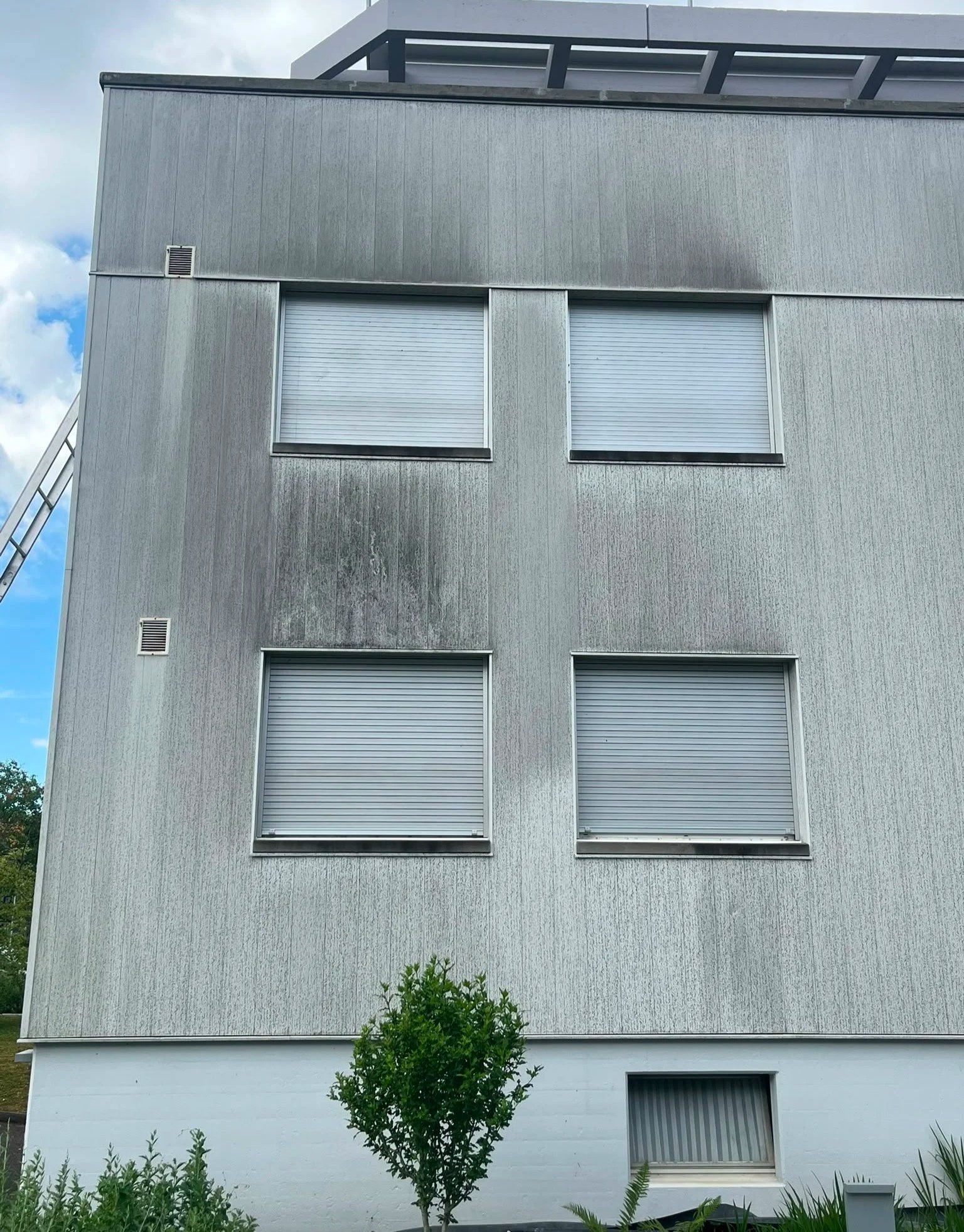Mehrstöckiges Gebäude mit geschlossenen Fensterläden, grauer Fassade und einem kleinen Baum vor dem Gebäude, im Hintergrund teilweise blauer Himmel mit Wolken.