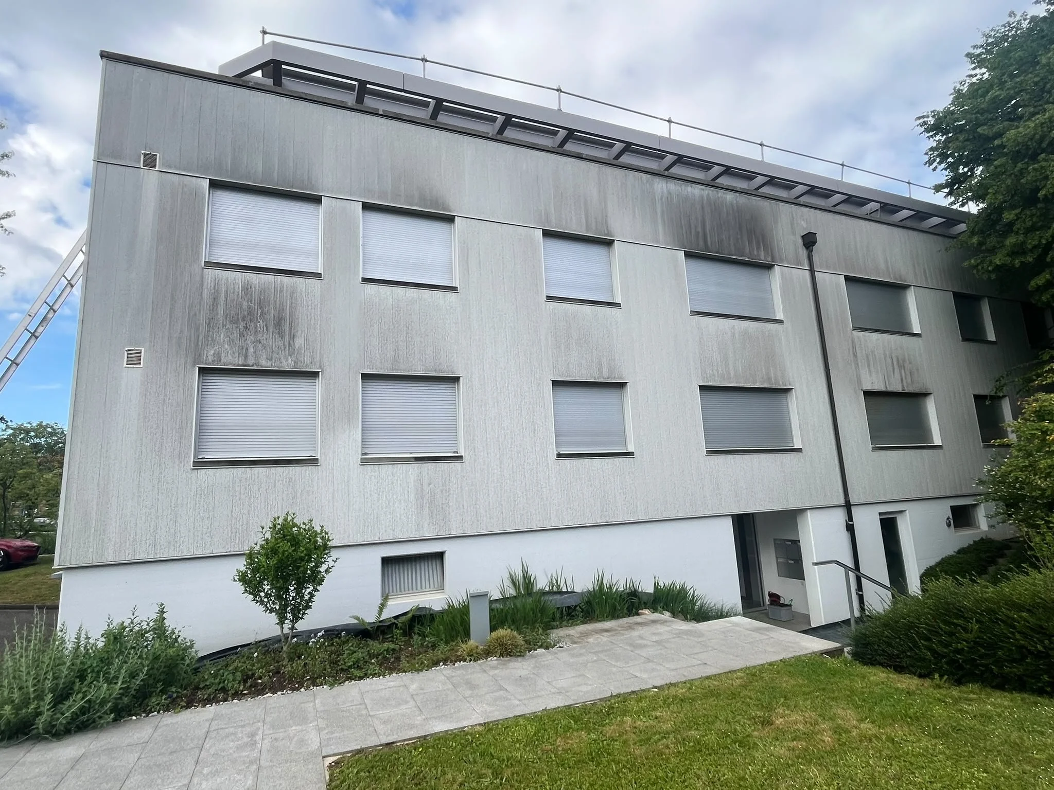 Modernes vierstöckiges Gebäude mit grauer Fassade und mehreren geschlossenen Rollläden. Es gibt einen Eingangsbereich mit Treppen, umgeben von einem gepflegten Garten mit Büschen und einer Pflanze. Die Straße im Vordergrund ist gepflastert, der Himmel ist bewölkt.