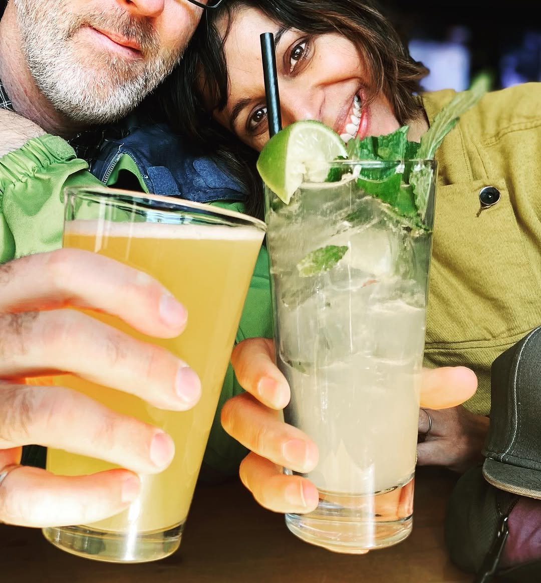 Two people smiling with drinks in a bar or restaurant setting, one holding a yellow-colored drink and the other holding a mojito with lime and mint.
