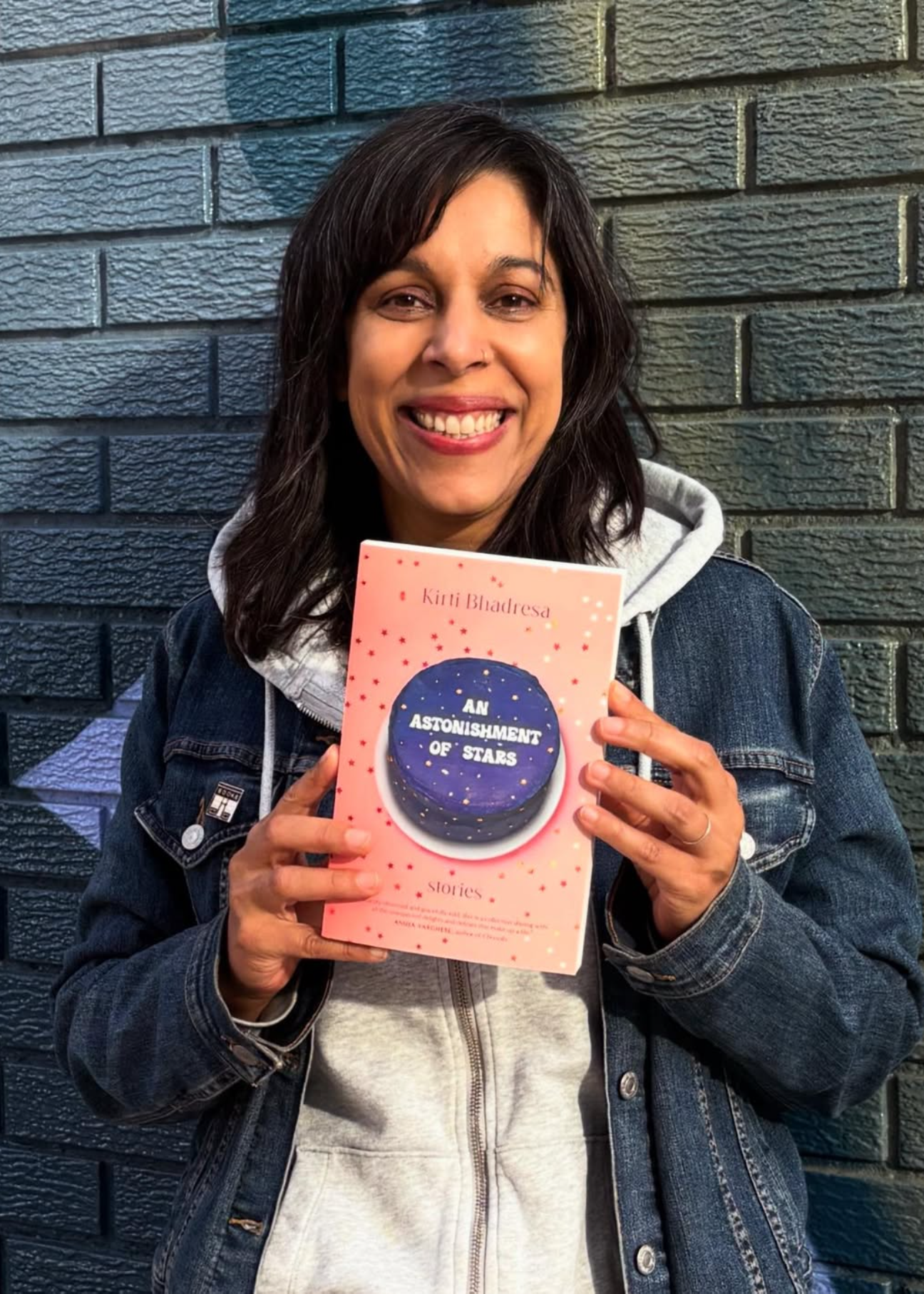 A woman with dark hair and a denim jacket is smiling and holding a pink book titled "An Astonishment of Stars" in front of a dark brick wall.