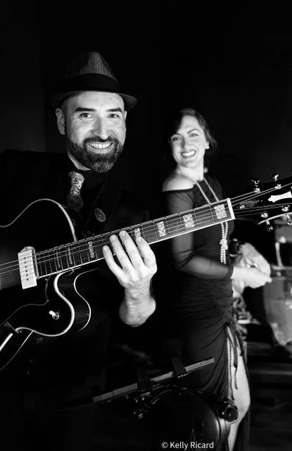 Man playing guitar, woman smiling in background at a musical performance