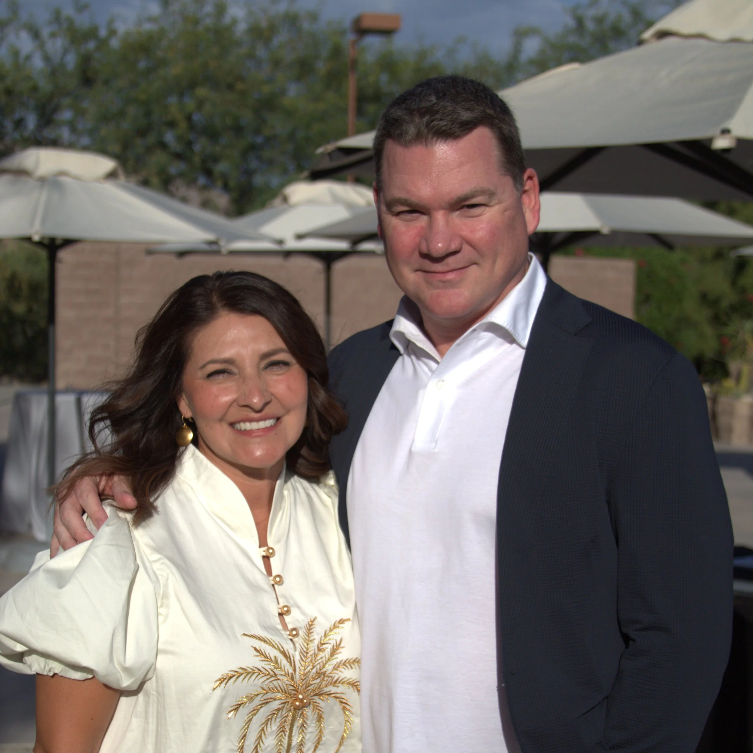 Partial view of a woman with dark brown hair smiling and wearing a white blouse with gold embroidery, and a man in a dark suit with a white shirt, standing outdoors near white umbrellas with greenery and a cloudy sky in the background.