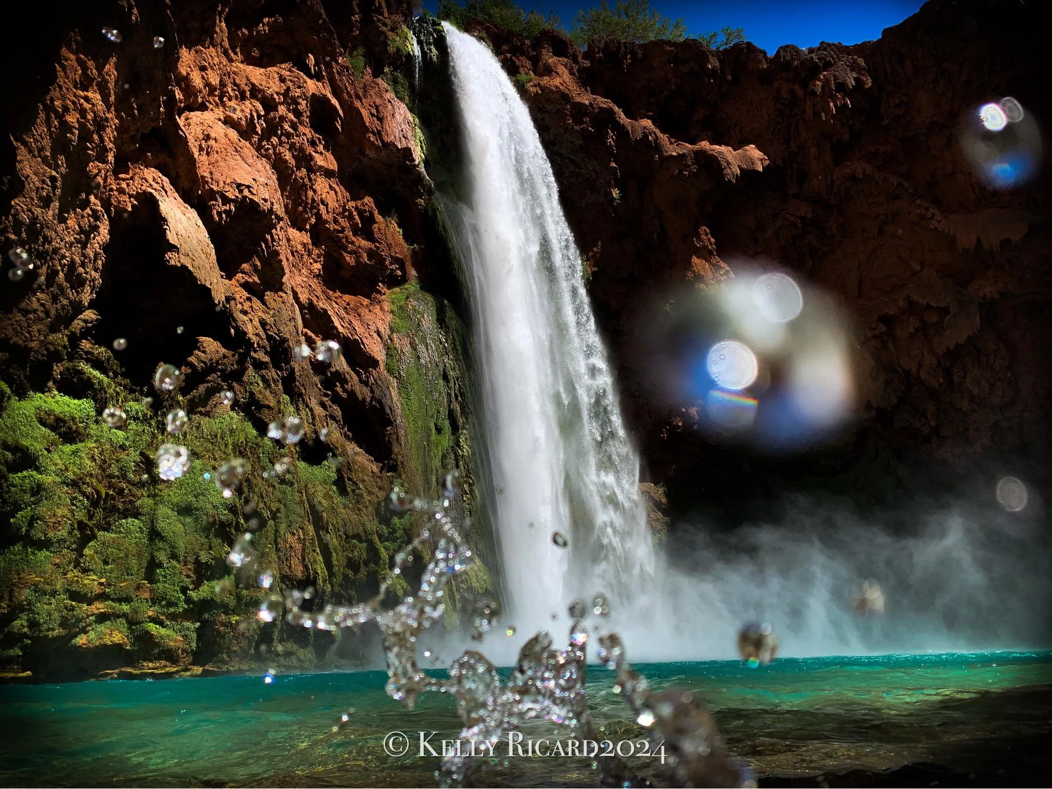 A tall waterfall flowing over reddish-brown rocks into a turquoise pool below, with sunlight creating reflections on the water and some green moss on the rocks.