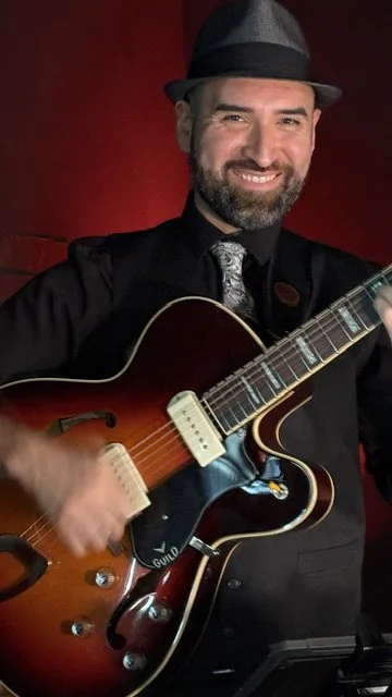 A man with a beard and a hat smiling while holding an electric guitar, standing against a red background.