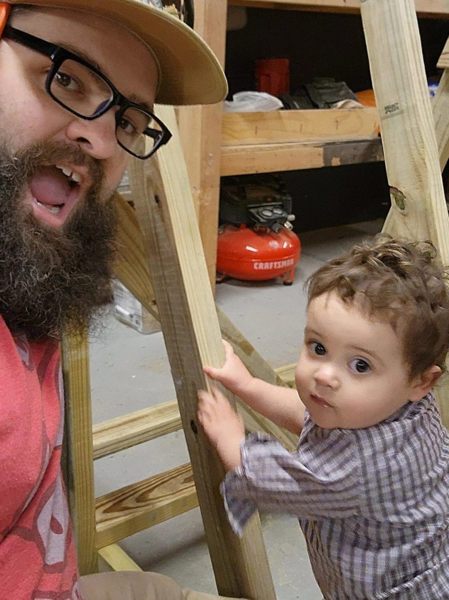 A bearded man wearing glasses and a tan hat is smiling while interacting with a young boy who has curly hair and is wearing a plaid shirt. They are surrounded by wooden frames, possibly at a construction or woodworking site.