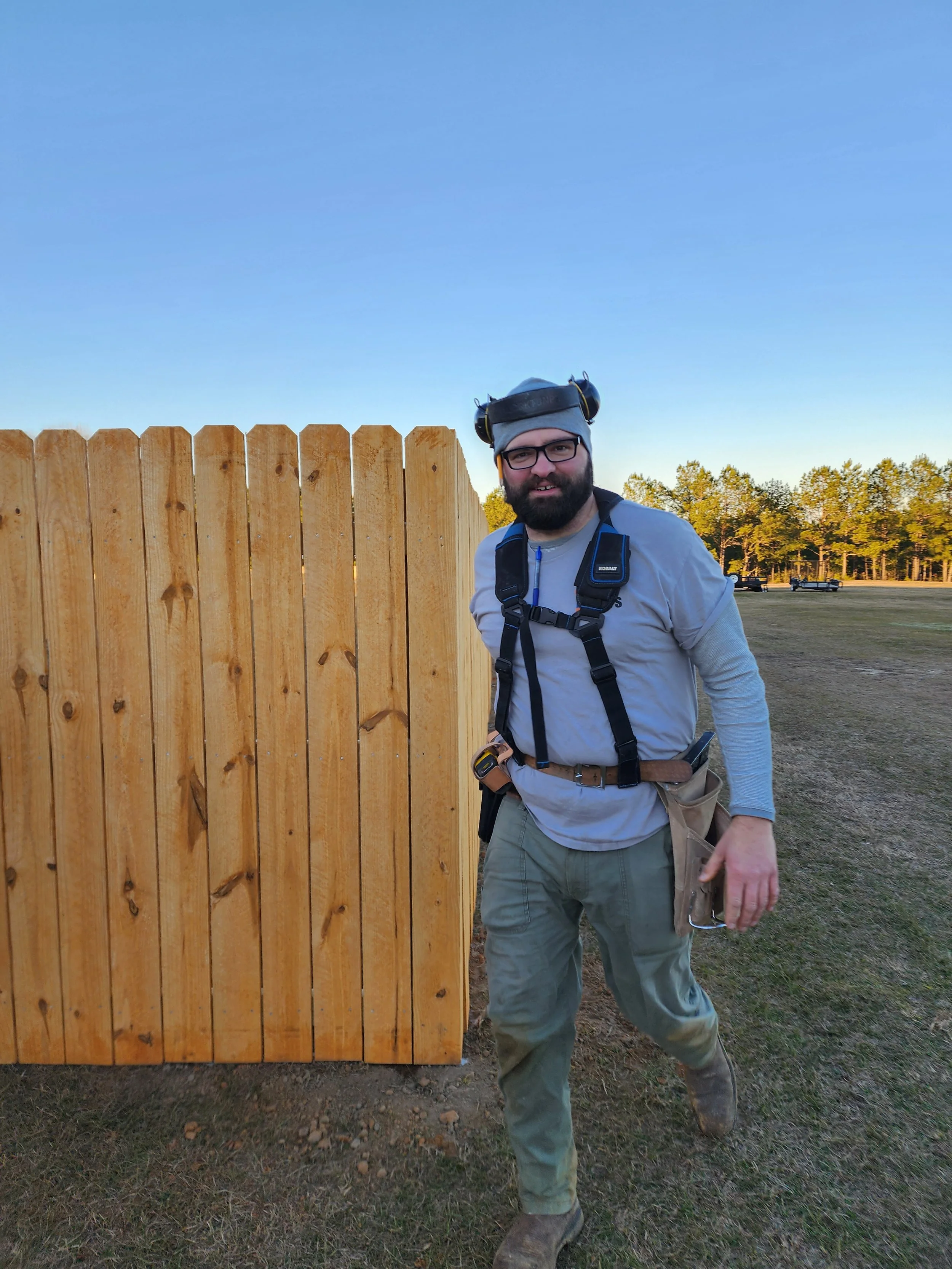 A man with a beard and glasses standing next to a tall wooden fence, wearing work clothes, gloves, and a protective helmet with ear protection, outdoors on a grassy area with trees in the background.