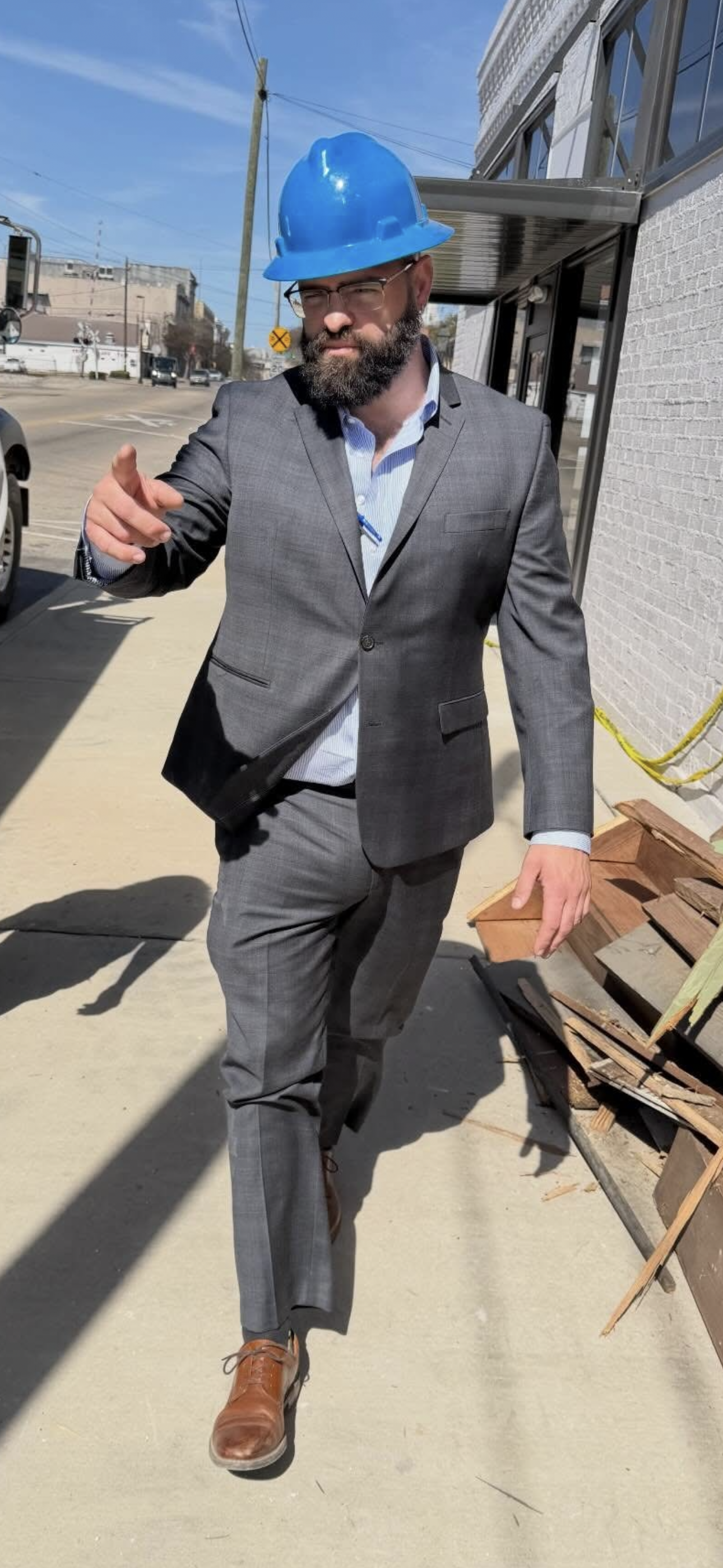 A man wearing a gray business suit, brown shoes, and a blue construction helmet walking outdoors on a sunny day, pointing toward the camera with a serious expression.