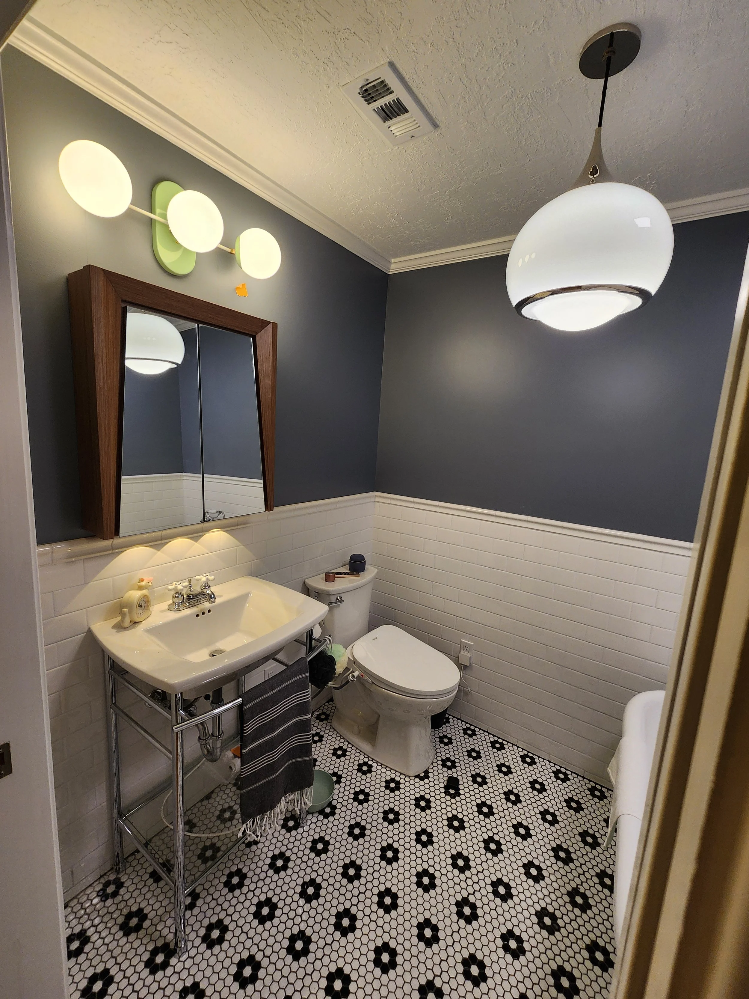A small bathroom with a black and white hexagonal tile floor, a white porcelain sink on a metal stand, a toilet, a mirror with a wooden frame, and navy blue walls. There is a modern ceiling light fixture and a wall-mounted light fixture with three round frosted glass shades.