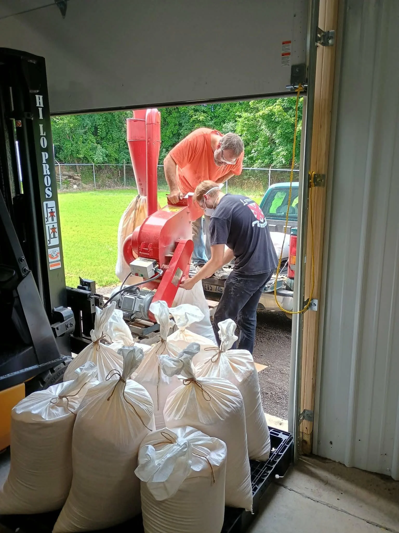 Two people working with a red machine outside, near large white bags filled with an unknown material, in a yard with a green lawn and trees, viewed from inside a garage or workshop.