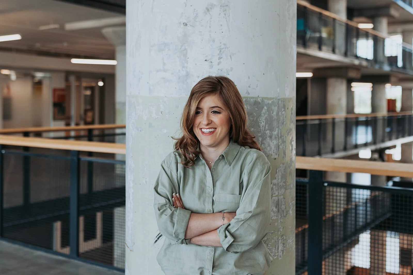 A woman with shoulder-length brown hair, wearing a light green button-up shirt, leaning against a concrete pillar with her arms crossed and smiling, in a modern office or industrial space.