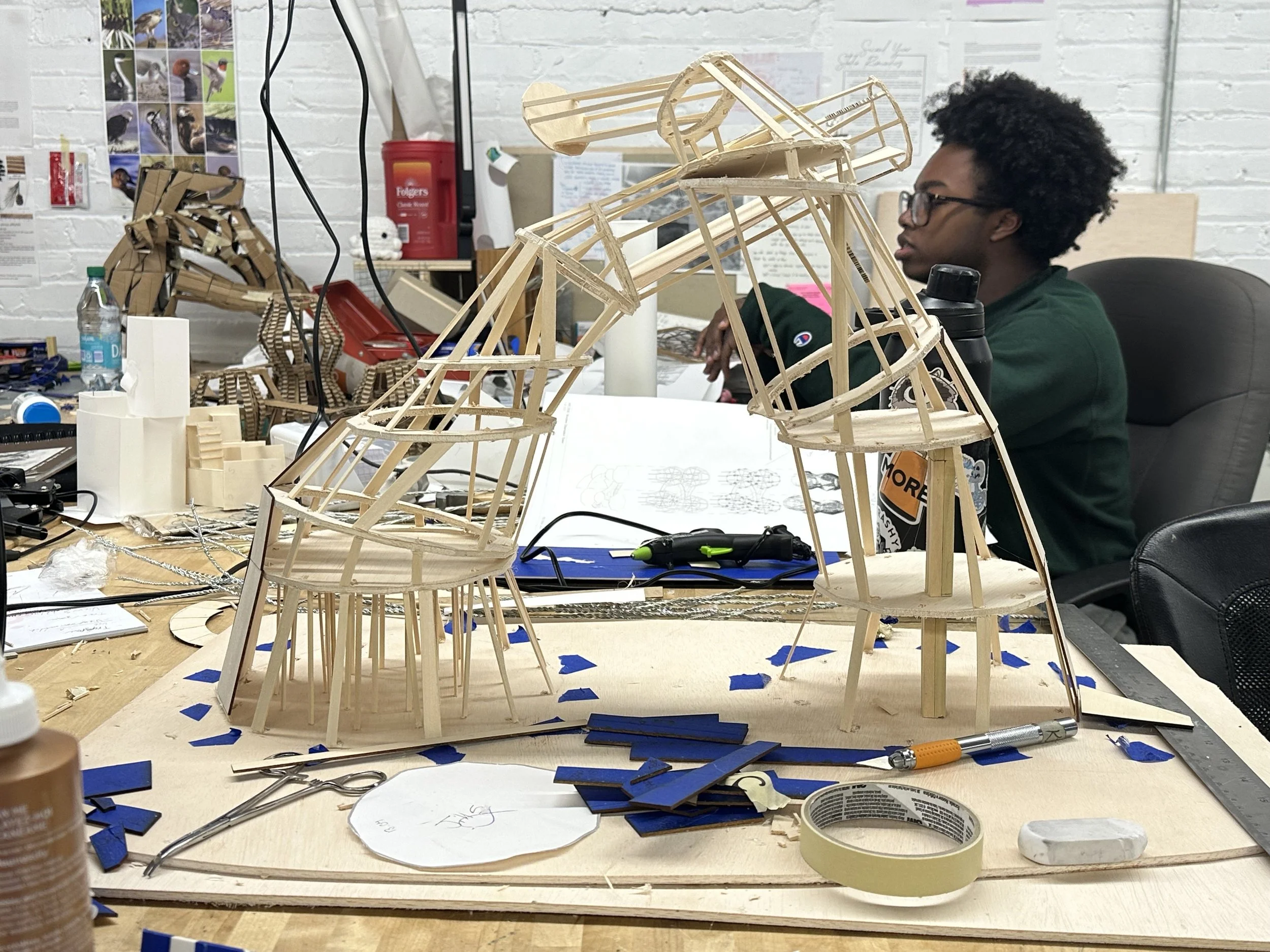 A person with glasses and curly hair sits at a cluttered workspace working on a large wooden model, possibly an aircraft or spaceship, with other models and tools on the table around them.