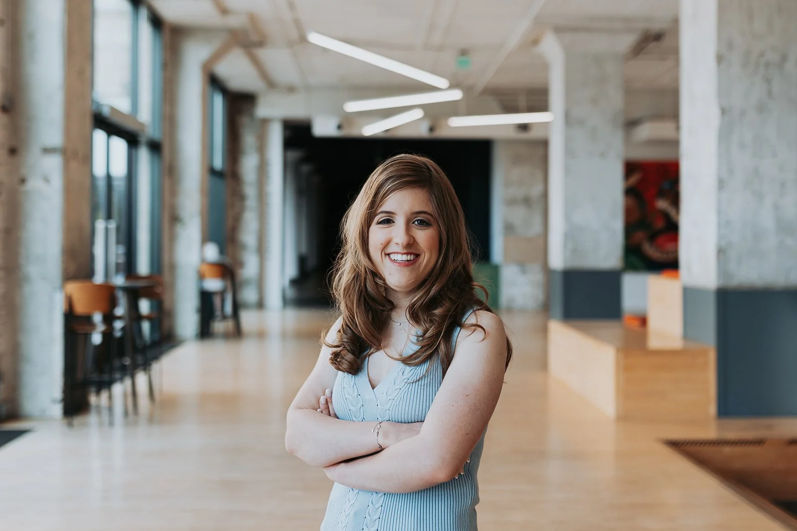 A young woman with long wavy brown hair and a light blue sleeveless dress smiling and crossing her arms in a modern indoor space with concrete walls, large windows, and contemporary lighting.