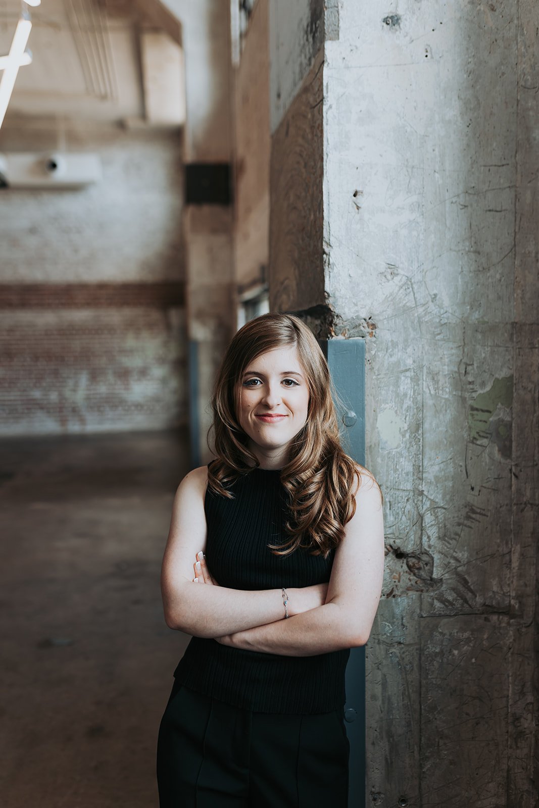 A young woman with wavy brown hair, wearing a sleeveless black top and black pants, standing with arms crossed against a textured concrete wall in an industrial-style interior.