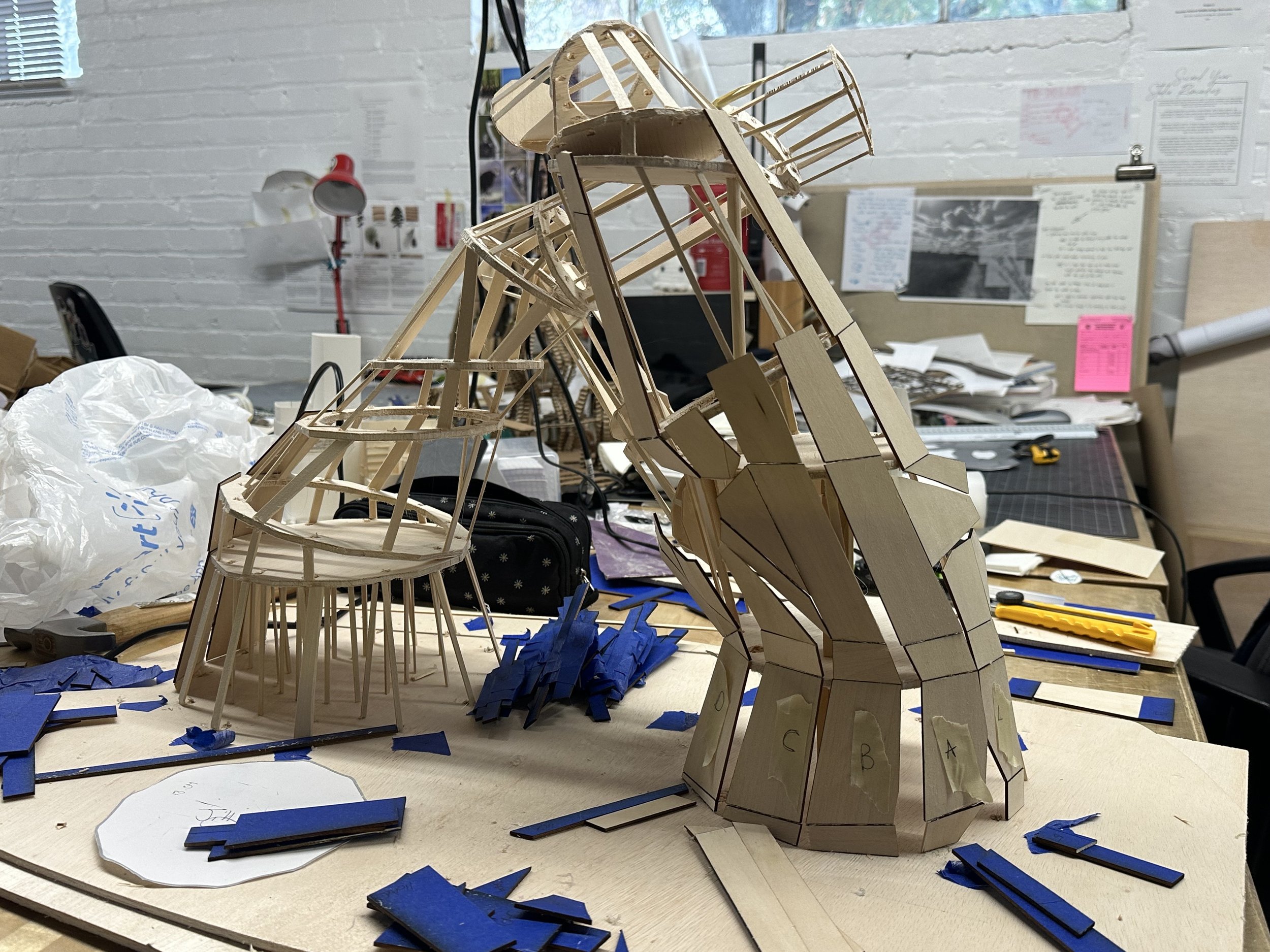 A detailed wooden model of a giraffe being built on a table with scattered blue foam pieces and paper notes surrounding it. The background shows a workshop with white brick walls, office supplies, and informational posters.