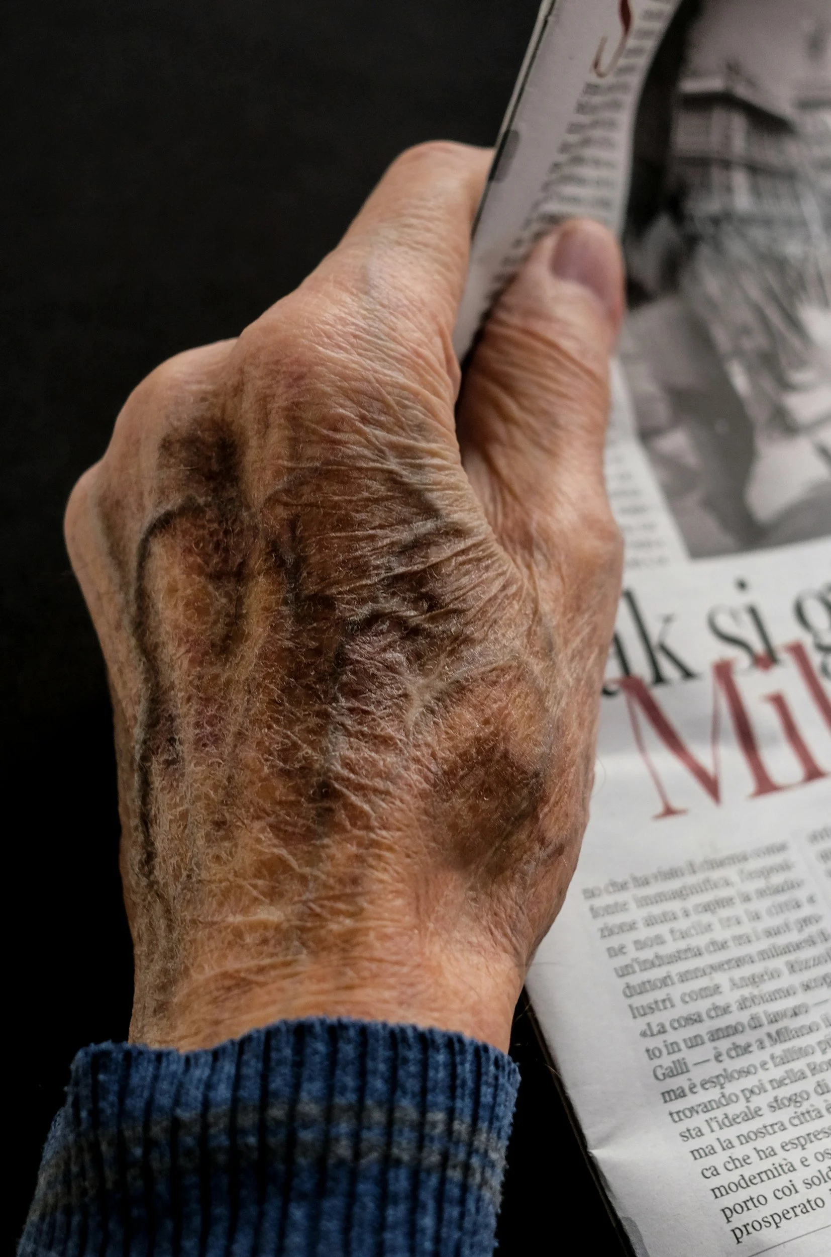 Close-up of an elderly person's hand holding a paper with text, wearing a blue ribbed sweater.