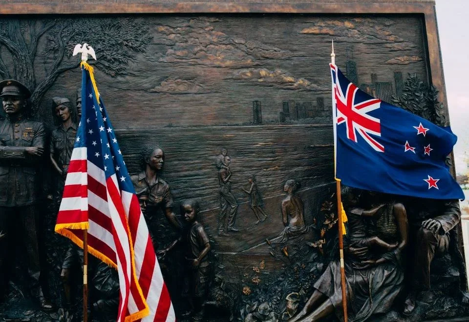 A bronze memorial wall with a relief depicting a family scene, flanked by an American flag and a New Zealand flag.