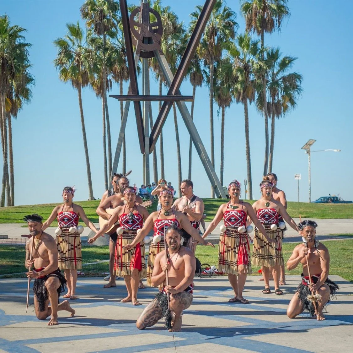 Group of indigenous performers in traditional attire performing a dance outdoors, with a clock tower and palm trees in the background.