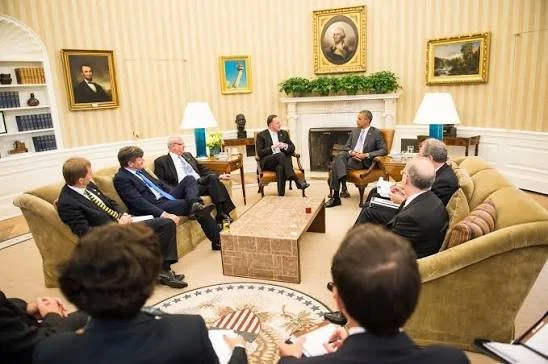 Group of people in a formal meeting in the Oval Office, with portraits and a fireplace in the background.