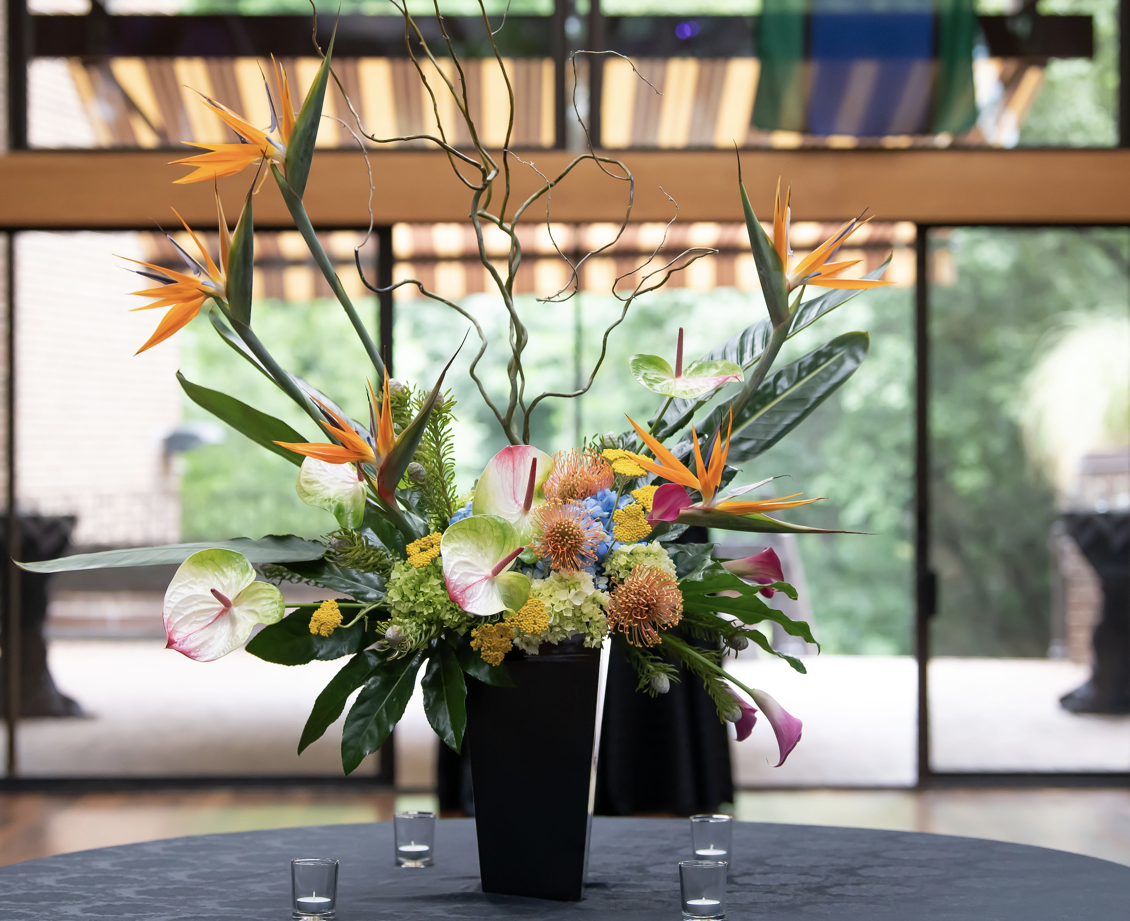 A floral arrangement with orange, pink, yellow, and green flowers and leaves, placed in a black vase, on a table with small clear candle holders in front of it, in front of large windows with a view of trees outside.