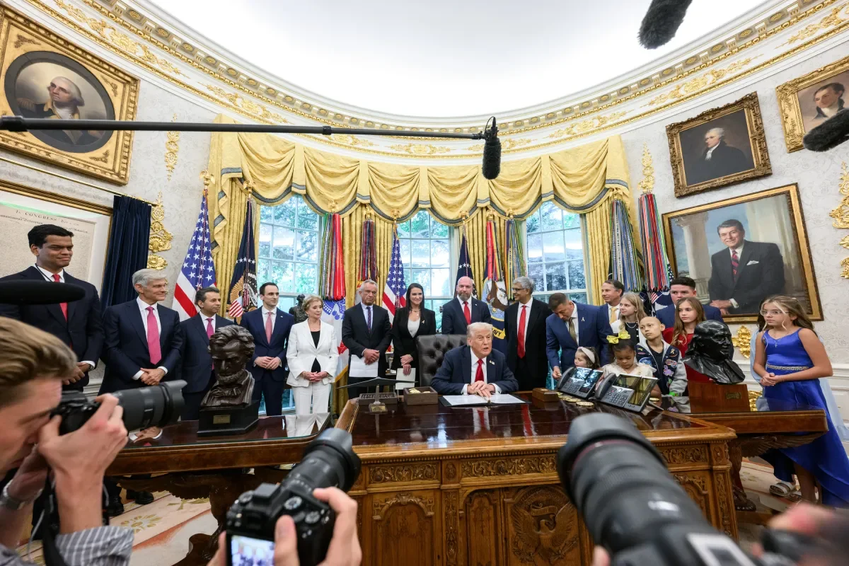 Former President Donald Trump sitting at the Resolute Desk in the Oval Office, surrounded by people including adults and children, with cameras capturing the moment. The room has gold drapes, American flags, and portraits on the walls.