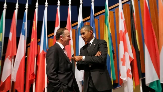 Two men in suits talking and smiling in front of a background of multiple international flags.
