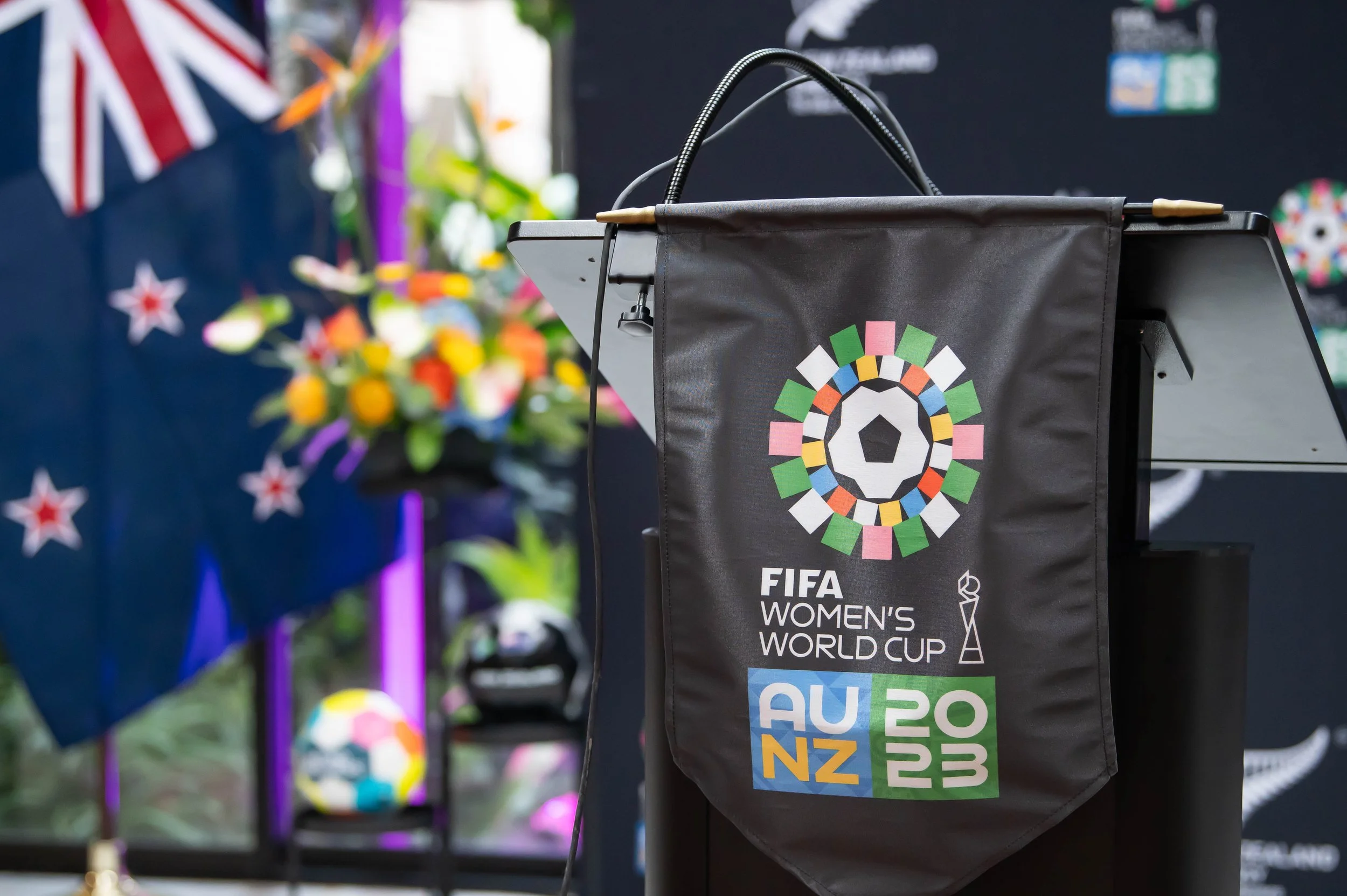 A podium with the FIFA Women's World Cup logo, decorated with the flags of different countries, and a colorful background with flowers and a display of soccer balls.