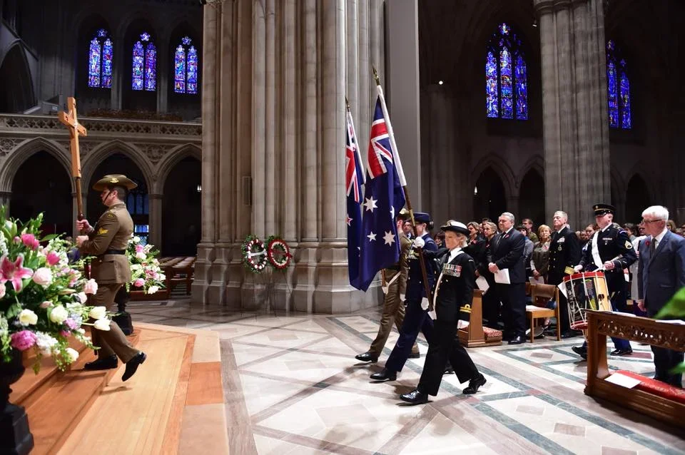 Military procession inside a church during a memorial, with soldiers carrying flags and a person holding a large wooden cross, surrounded by attendees and floral wreaths.