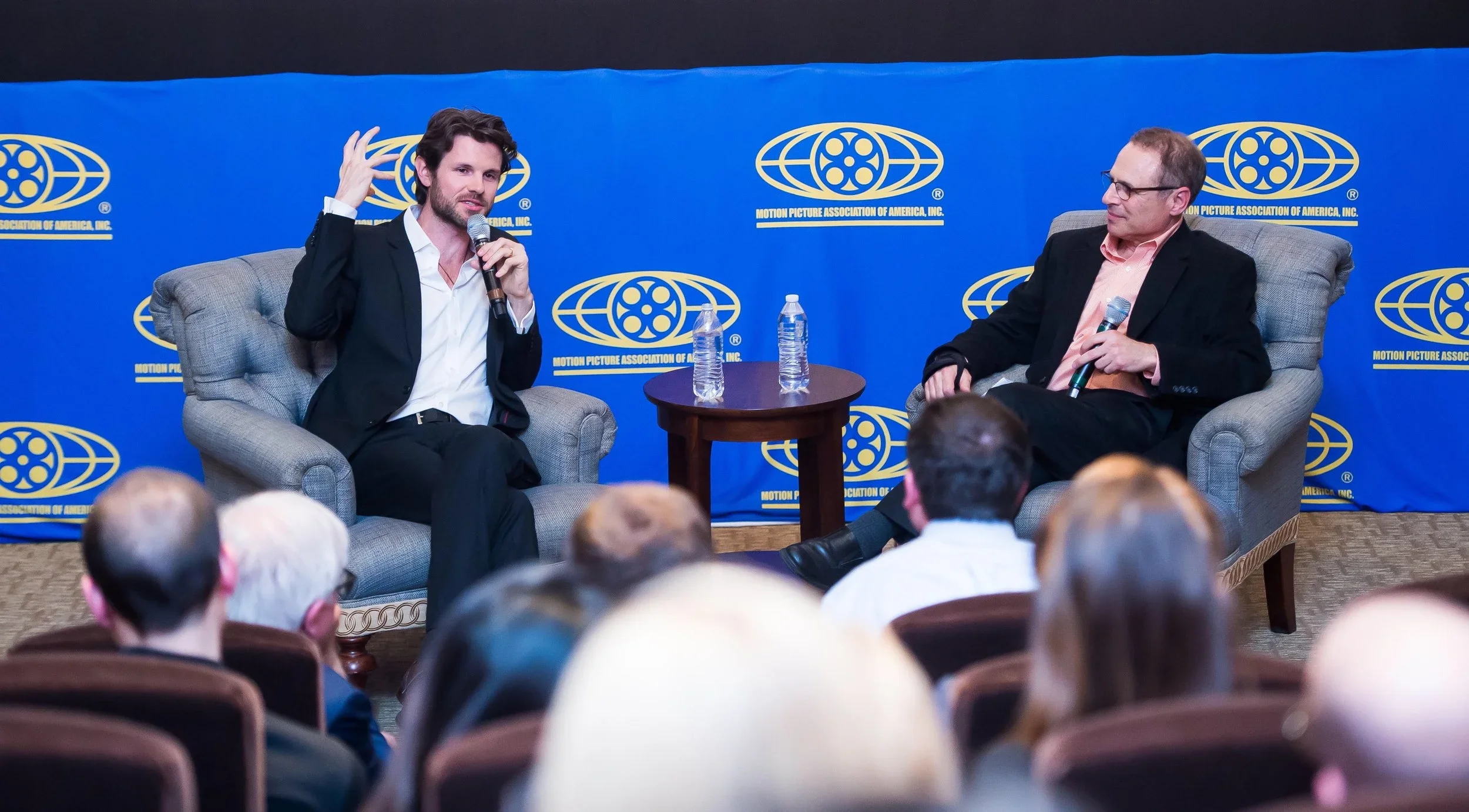 Two men are seated on stage at a panel discussion or interview, with a blue backdrop displaying the logo of the Motion Picture Association of America, Inc. The man on the left is talking into a handheld microphone, while the man on the right holds a microphone in his lap and listens. Two water bottles are on a small round table between them, and audience members are visible in the foreground.