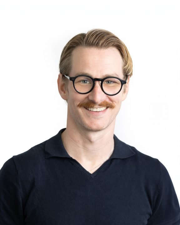 A man with light skin, blonde hair, glasses, and a mustache smiling against a plain white background.