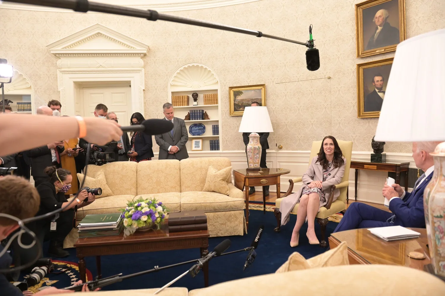A woman sitting on a yellow armchair in the Oval Office, smiling, with people filming her and taking photos around her.