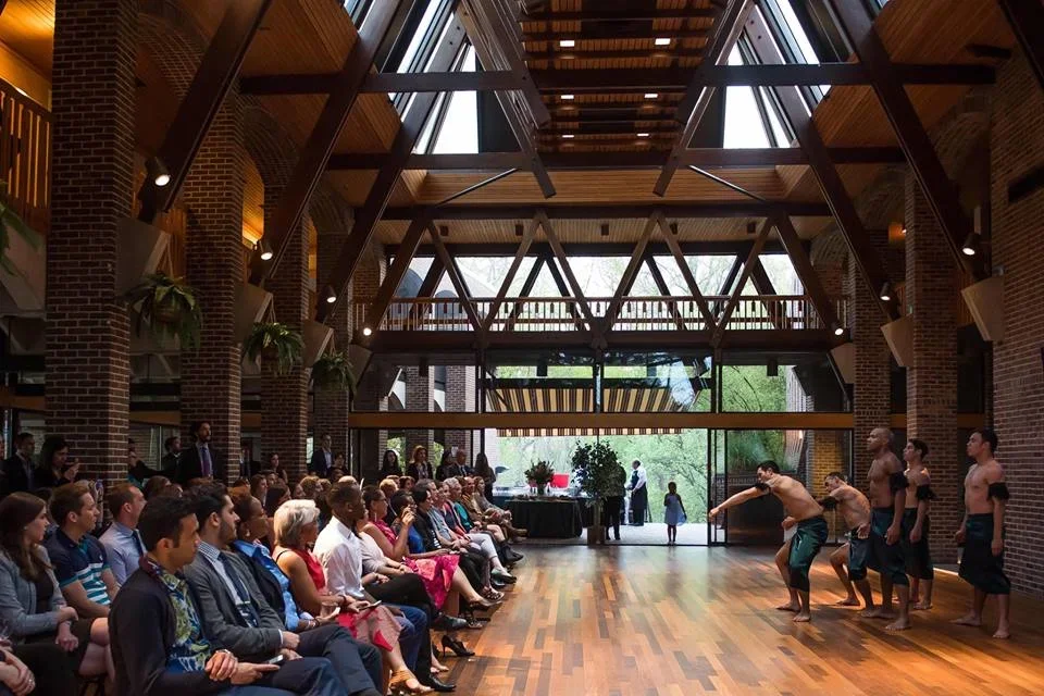 Group of performers doing a traditional dance in a spacious, modern hall with a high wooden ceiling and brick walls, attended by an audience seated on both sides.