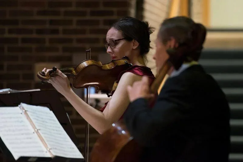 Two women playing violin and cello during a performance in a dimly lit room.
