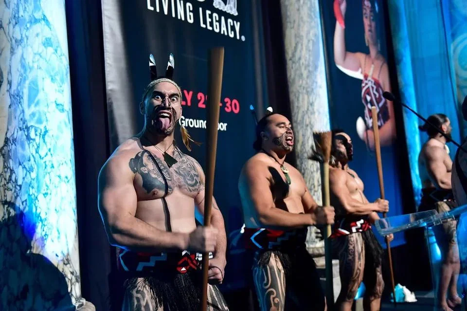 Group of men on stage dressed in traditional Polynesian costumes, holding spears, with face paint and tribal tattoos, performing a cultural dance.