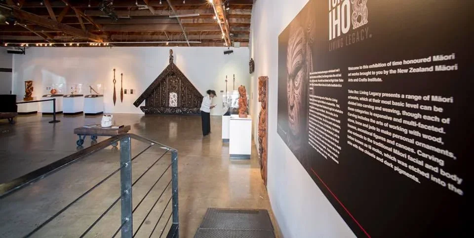 An art exhibition in a gallery featuring Māori arts, with display cases and traditional carved wooden artwork, and informational panels about Māori culture.