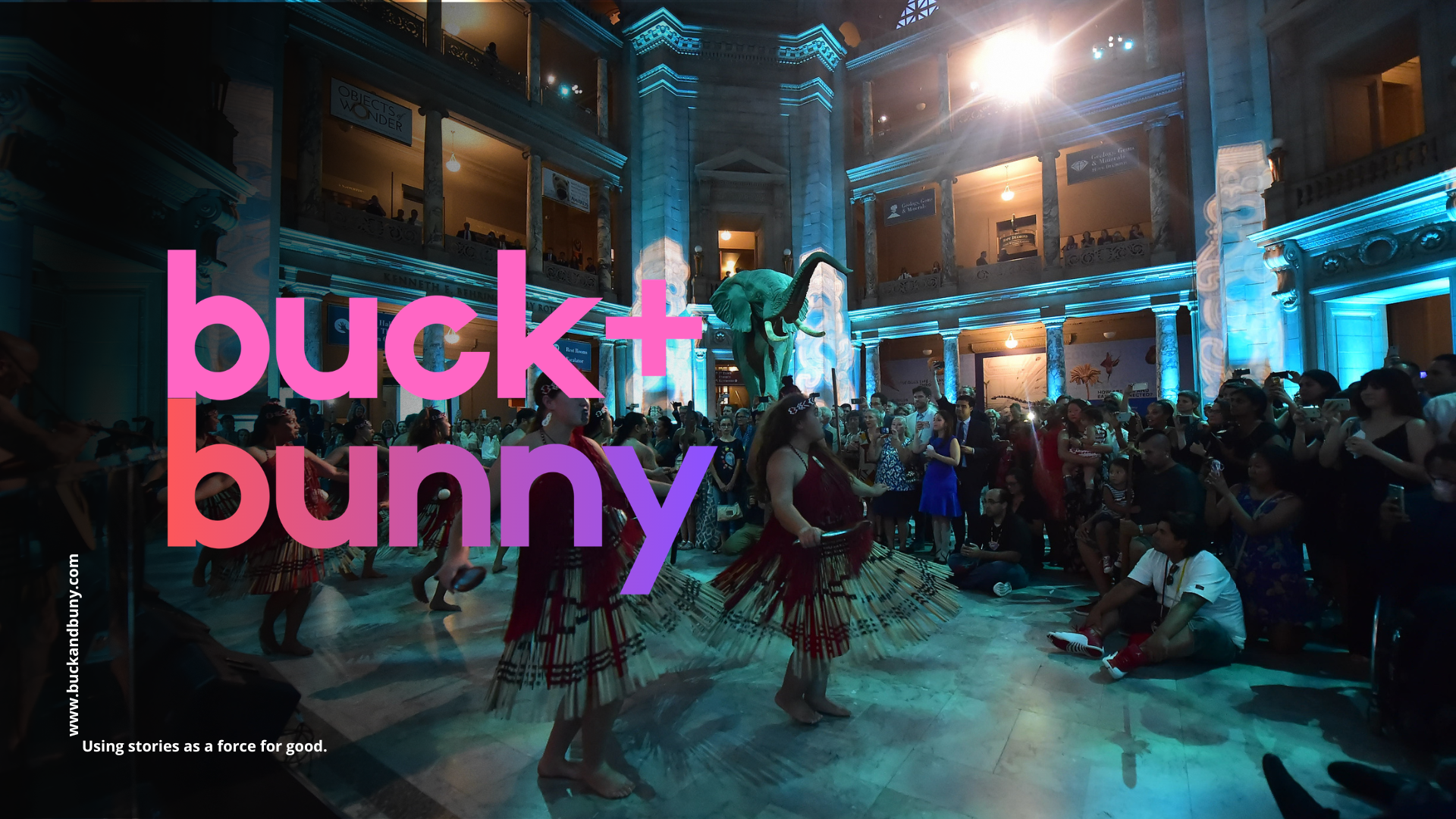 A lively indoor event at the American Museum of Natural History, featuring indigenous dancers in traditional attire performing in front of a large crowd. The museum's architecture with columns and exhibits can be seen in the background, with a prominent dinosaur model and ambient blue lighting.