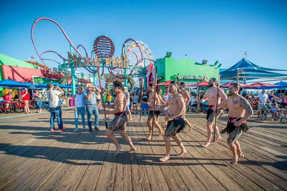 Group of shirtless men in traditional Polynesian skirts dancing on a wooden pier at an amusement park with rides and people in the background.