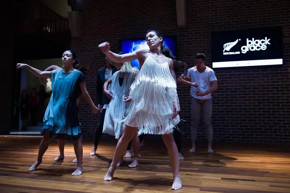Group of dancers performing on wooden stage in front of a brick wall, with a Black Grace logo on a screen in the background.