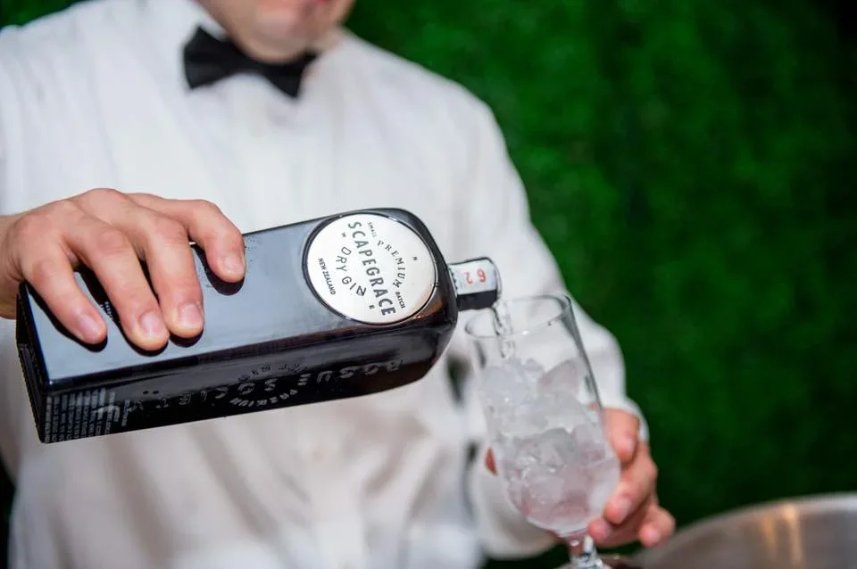 A bartender in a white shirt and black bow tie pours Top Shelf Scotch from a black bottle into a glass filled with ice, against a blurred green background.