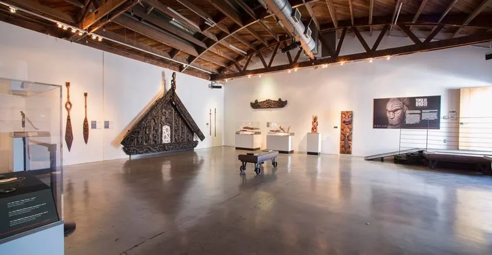 An art gallery with various sculptures and artifacts displayed on white walls and pedestals, featuring tribal and historical pieces, including masks, carved figures, and weapons, with a polished concrete floor and an exposed wooden beam ceiling.
