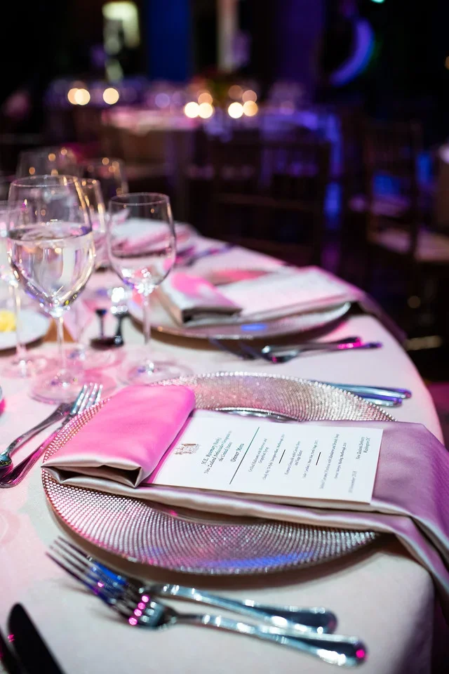 Elegant banquet table set for a formal event with wine glasses, silverware, pink napkins, and a menu, decorated with a shimmering placemat, in a dimly lit venue with purple and blue lighting.