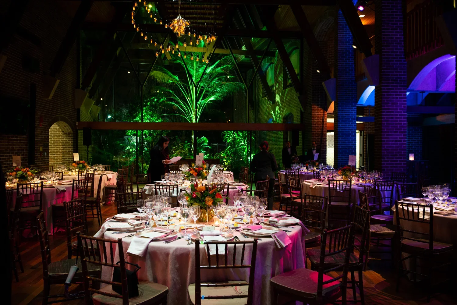 An elegant event space with round tables covered in white tablecloths, set with glassware, plates, and floral centerpieces. Behind the tables, large windows reveal a lush, green tropical garden illuminated with vibrant green lighting. The high ceiling has colorful accent lighting and chandeliers, with brick walls and wooden accents.