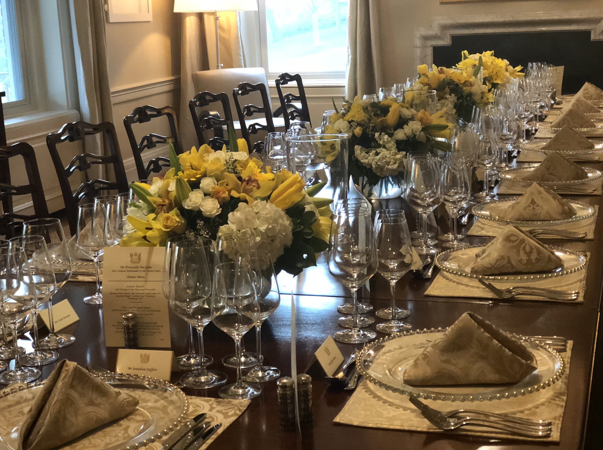 A formal dining table set with multiple glasses, silverware, gold trimmed plates, beige napkins, and elegant floral centerpieces with yellow and white flowers in a well-lit room with large windows and a fireplace