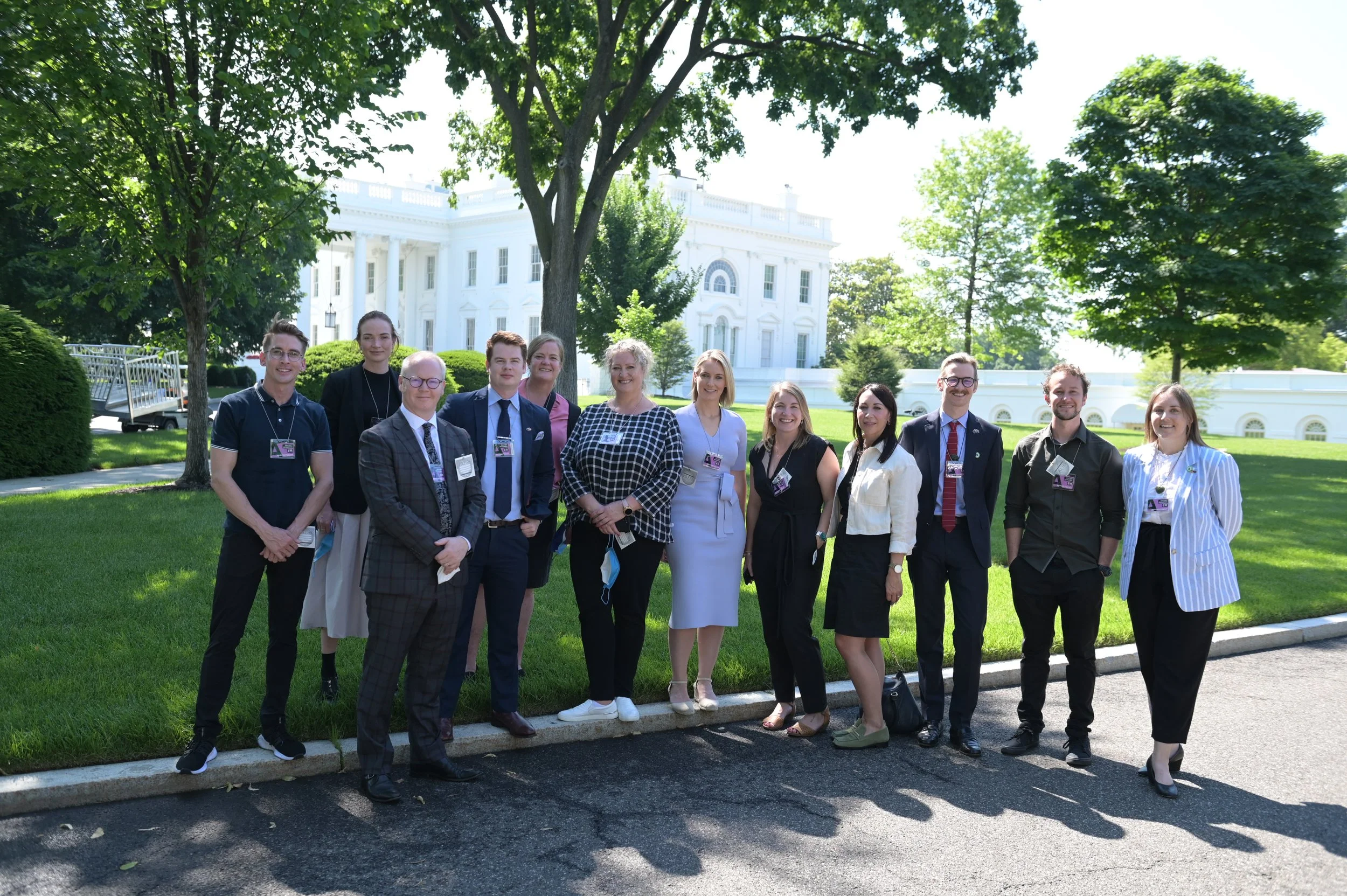 Group of professionally dressed people standing in a row outdoors in front of the White House, smiling for a photo on a sunny day with lush green trees and grass.