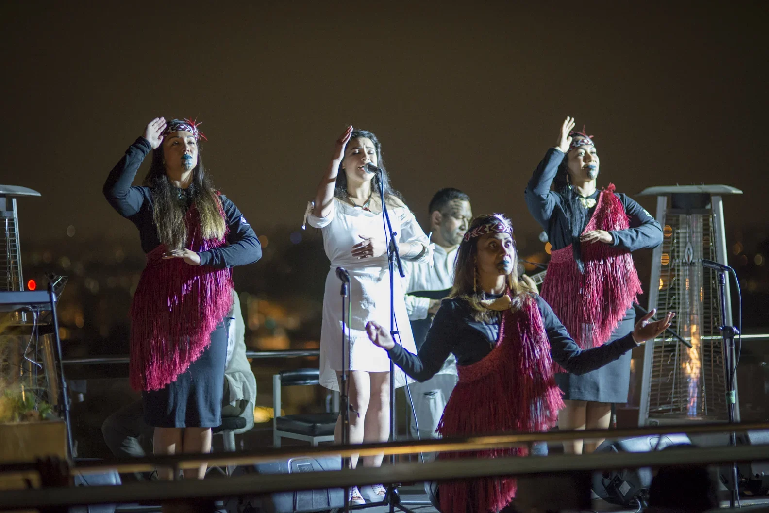 Group of five women singing and performing on a rooftop at night, with city lights in the background. Some women are dressed in traditional attire with red fringed scarves and tribal headbands.