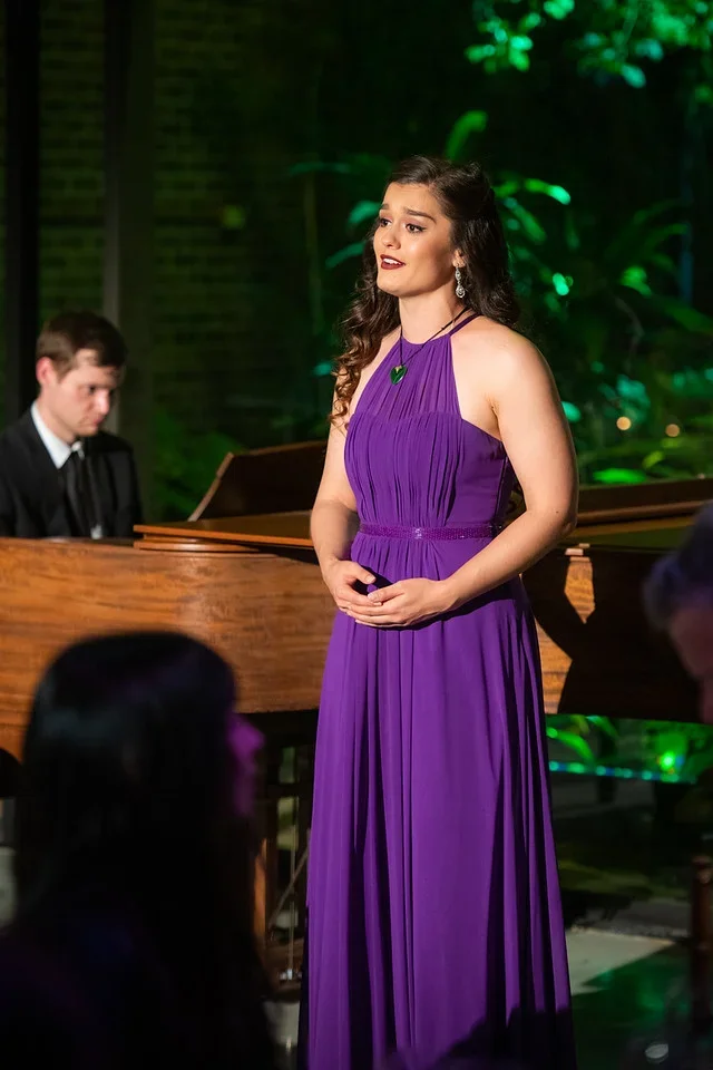 A woman in a purple dress singing on stage with a pianist playing in the background at a concert or performance venue.